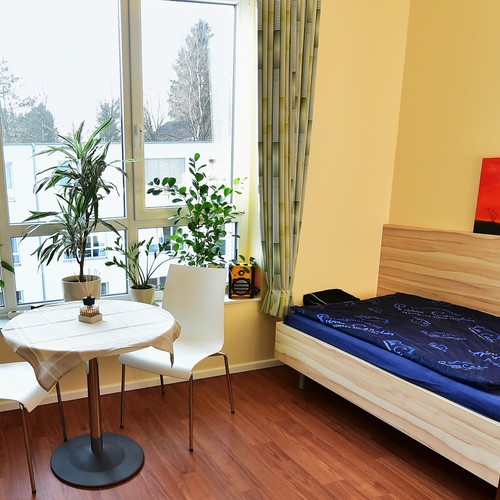 Beispielhaft ein helles Einzelzimmer mit Bett, Sitzgelegenheit und großen Fenstern.
